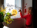 Child painting pumpkin on window preparing celebrate Halloween. Little kid draws decorates room interior with paper bats