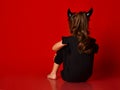 Child with devil horns, ponytail, in black blouse and leggings, barefoot. She sitting against red background. Back view. Close up Royalty Free Stock Photo
