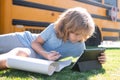 Child does school homework laying on grass in the park near school bus. School kid outdoor.