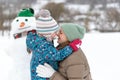 Child embracing mom against the snowman wearing hat and scarf. Portrait of mother and child in winter outside