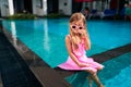 Little girl in pink dress, heart-shaped sunglasses sits at pool edge, feet dipped in water. Child enjoys summer day Royalty Free Stock Photo