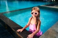 Smiling girl with pink swimsuit, sunglasses sits at pool edge summer day. Child enjoys sunny weather, refreshing in blue Royalty Free Stock Photo