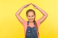 Child feel safe. Portrait of happy little girl with braid in denim overalls standing under house roof gesture