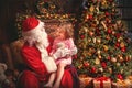 child girl in nightgown sitting on lap of Santa Claus around Christmas tree