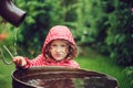 Child girl in red raincoat playing with water barrel in rainy summer garden. Water economy and nature care Royalty Free Stock Photo