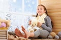 Child girl is sitting on a window sill and playing with bear toy. Beautiful view outside the window - sunny day in winter and snow