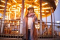 Child girl is standing at the street against background of carousel and evening city lights. Royalty Free Stock Photo