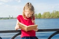 Child girl student wearing glasses with backpack is reading book. Urban background, river, sky. Royalty Free Stock Photo