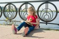 Child girl student wearing glasses with backpack is reading book. Urban background, river, sky Royalty Free Stock Photo