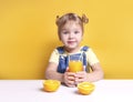 Child girl at table with glass of orange juice.Fresh fruits,vitamin nutrition. Caucasian kid portrait with beverage on yellow