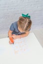 Child, a girl at the table writes, draws on a piece of paper, against a white brick wall Royalty Free Stock Photo