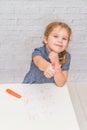 Child, a girl at the table writes, draws on a piece of paper, against a white brick wall Royalty Free Stock Photo