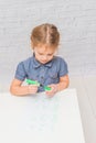 Child, a girl at the table writes, draws on a piece of paper, against a white brick wall Royalty Free Stock Photo