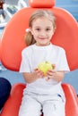 Child girl in white clothes at reception at a pediatric dentist smiles and laughs. little girl is holding a big green apple in her Royalty Free Stock Photo