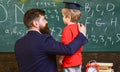 Child in graduate cap listening teacher, chalkboard on background, rear view. Teacher with beard, father teaches little son in