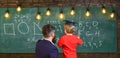 Child in graduate cap listening teacher, chalkboard on background, rear view. Instructive conversation concept. Teacher