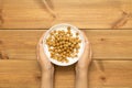 Child hands holding full bowl of yogurt with cereal balls on wooden table, top view. Quick and healthy food