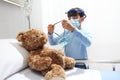 Child in the hospital puts oxygen mask on teddy bear on bed, wearing protective visor and surgical mask, corona virus covid 19 Royalty Free Stock Photo