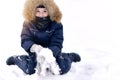 A child in a jumpsuit with a balaclava of a gaiter. Sitting on a winter day, a lot of snow with his hands in mittens