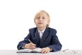 Child like boss sits at table and writes in notebook. Diligent student writes in copybook. Blond boy in jacket and shirt