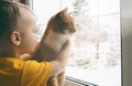 Child and pet at home. Red ginger tabby cat and little toddler boy looking out fo window watching falling snow. Domestic animal