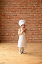 Child playing chef. Cute toddler girl in chef apron and hat eating sweet bun at brick wall in empty room Royalty Free Stock Photo