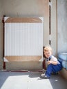 Adorable baby boy sitting near heating radiator in apartment.