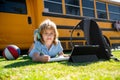 Child pupil does school homework laying on grass in the park near school bus. School kid outdoor.