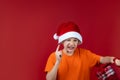 A child on a red background in a Santa Claus hat is holding a gift bag in his hands, grimacing and looking at him Royalty Free Stock Photo