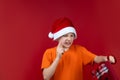 A child on a red background in a Santa Claus hat is holding a gift bag in his hands, grimacing and looking at him Royalty Free Stock Photo