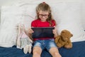 Child is resting at home. Little girl 6, 7 years old sitting in bed with toys, looking at her tablet, smiling Royalty Free Stock Photo