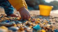 A child& x27;s hand picking up microplastics from the sand, with a bucket and shovel in the background, representing the
