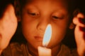 Child with sad eyes looks at flame of burning candle in dark. Memories or mourning for dead. Religious sacrament in church