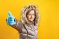 The child shows a thumb up. Happy cute little girl in a down jacket on a yellow background. Winter concept.