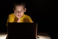 The child sits in the dark at the table and looks at the laptop