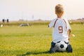 Child sitting on soccer ball on the football pitch. Kid in sports jersey uniform watching soccer training Royalty Free Stock Photo