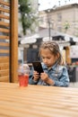 Child is sitting at table in street courtyard in cafe and looks intently at phone.