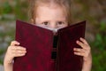 Child slyly narrowed his eyes and holds book. Little girl hiding behind book. Emotions of schoolgirl reading book Royalty Free Stock Photo