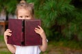 Child slyly narrowed his eyes, holds book. Little girl hiding behind book, looking at camera. Schoolgirl reading book Royalty Free Stock Photo