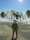 A child standing in the square in the sunshine in front of small gushing jets of water. Joint family vacation in the picturesque