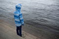 Child standing on stone steps near the water with his back in blue jacket with hood. Royalty Free Stock Photo