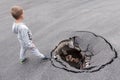 Child stands on brink of a large pit in asphalt. Danger of injury to children on city street. Road surface hole