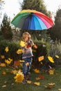 child stands under a rainbow umbrella, many colorful autumn leaves fall around