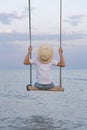 Child in straw hat riding on rope swing over water. Sea and blue sky background. Vertical frame Royalty Free Stock Photo