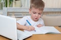 Cute little boy draws homework in a notebook sitting at the table in front of a laptop. Online home education concept Royalty Free Stock Photo