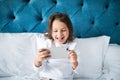 Child, technology and internet concept - little girl lying in bed with smartphone playing games at home