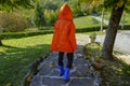 Child wearing orange raincoat, hood and blue rubby boots walking in autumn park. Rainy clothing