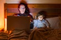 A child and a woman sitting on the bed watching the smartphone and the laptop