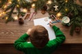 Child Writing Christmas Letter to Santa Claus. Top View of Kid sitting at Wooden Table with Xmas Tree Decoration Lights and Empty Royalty Free Stock Photo