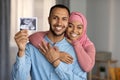 Childbirth concept. Happy black muslim couple expecting baby, showing sonography at camera